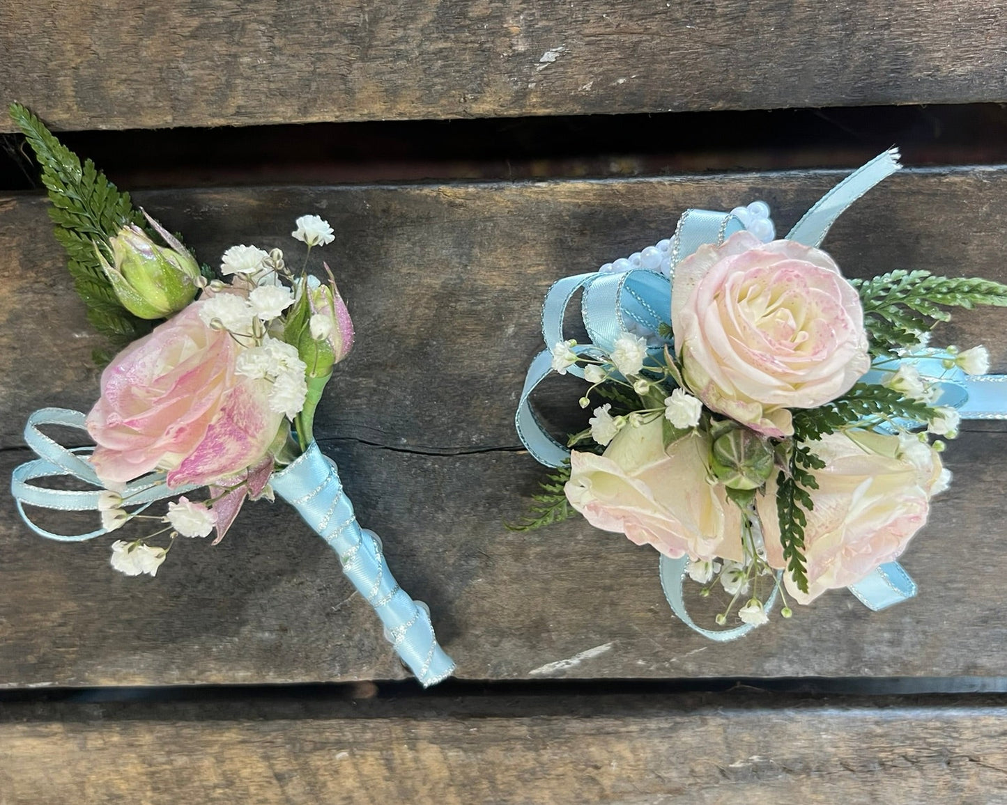 Two floral wrist corsages with pale pink and cream roses, baby's breath, green foliage, and light blue ribbons, displayed on a rustic dark wood plank background.