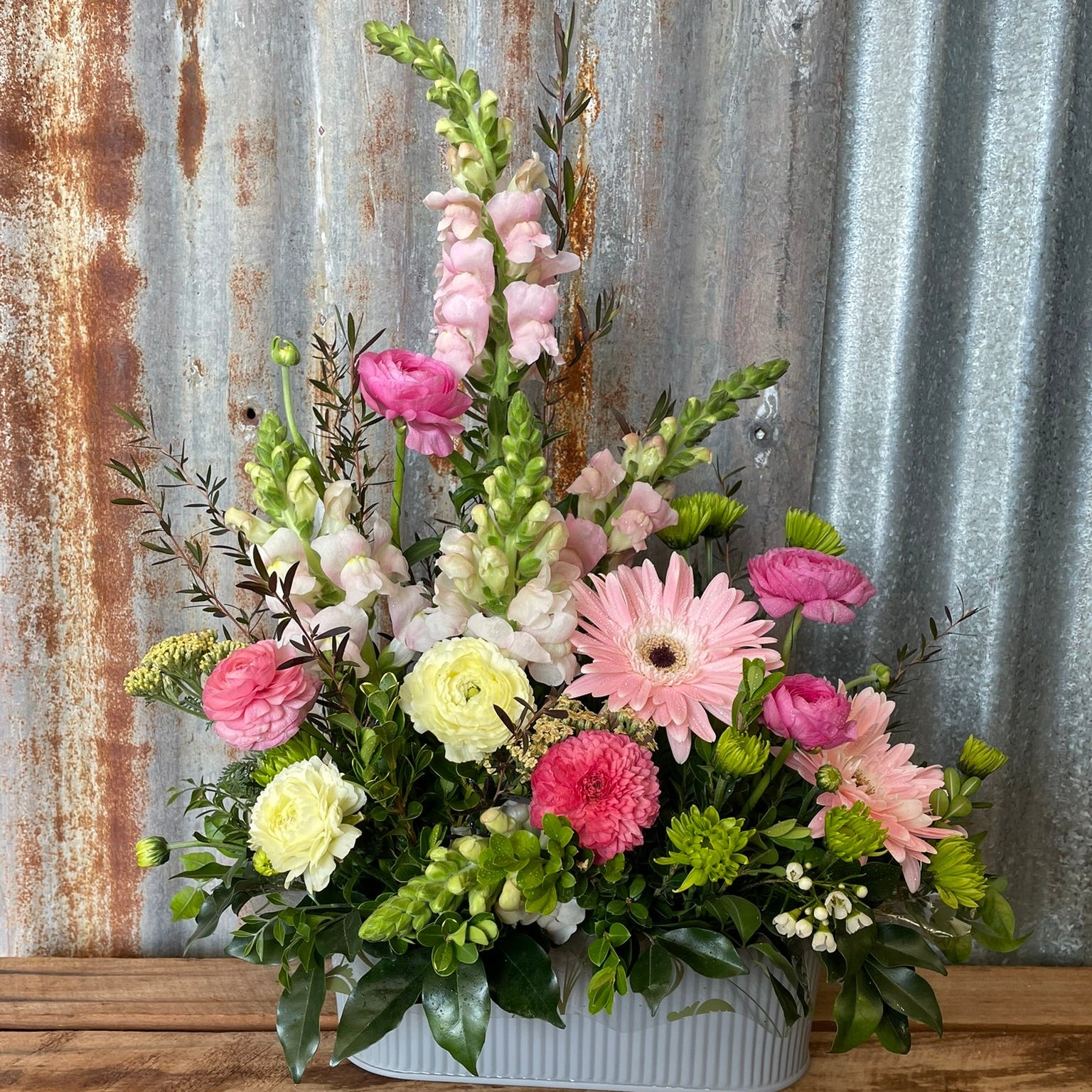 Floral arrangement with pink, white, and green flowers in a white container against a rustic metal and wooden background.