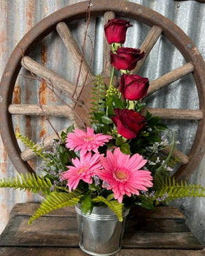Floral arrangement in a metal container on a wooden pallet with a rustic wheel in the background.
