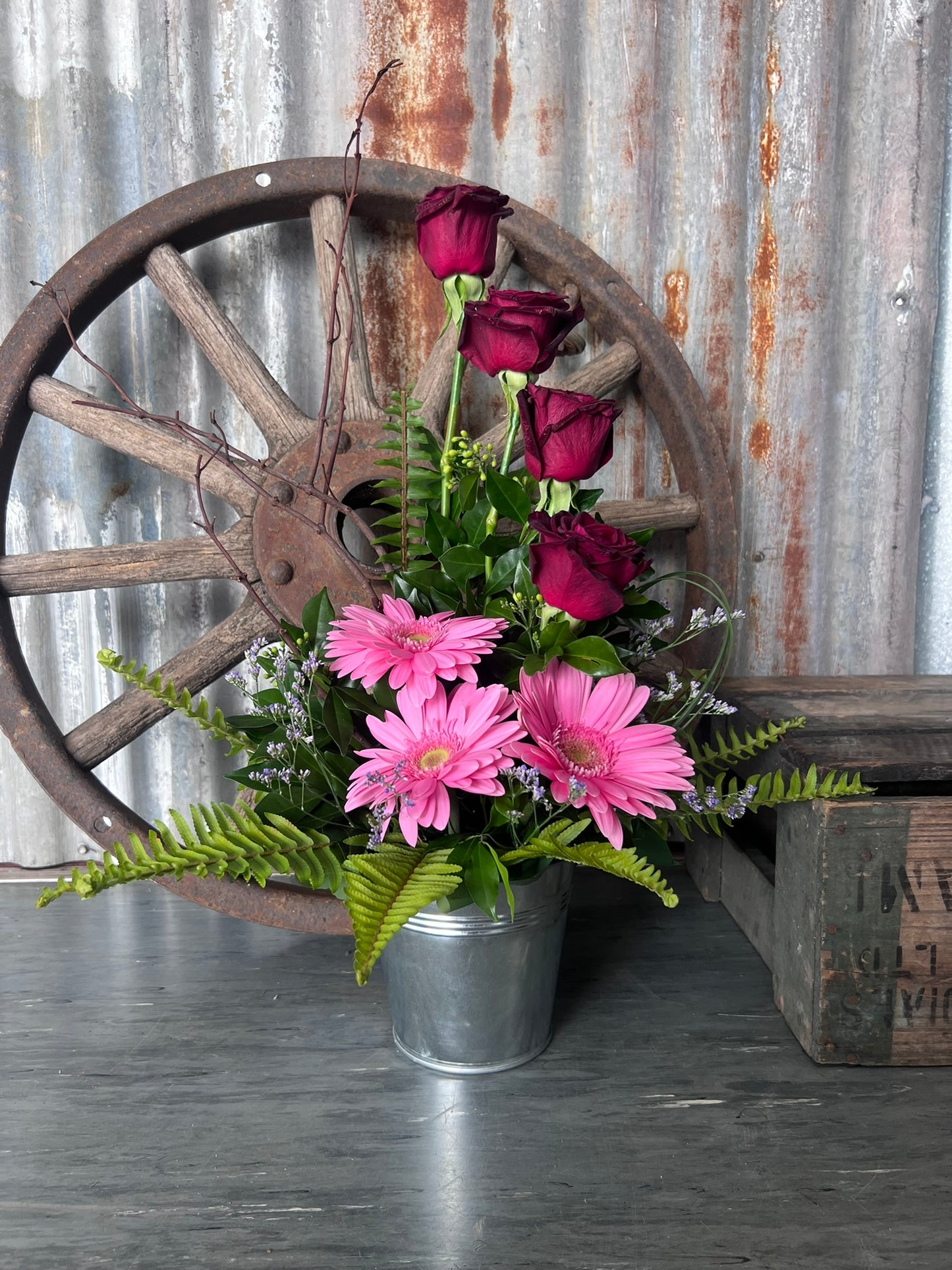 Floral arrangement in a metal container with a rustic wooden wagon wheel and wooden crate in the background.