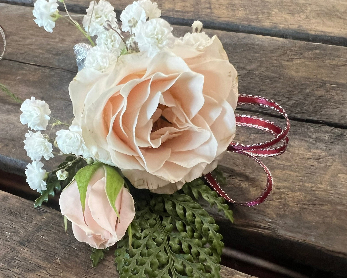 An artificial floral wrist corsage featuring a large pale peach-pink rose, a smaller matching rose bud, white baby's breath, and green fern leaves, tied with a red and white striped ribbon, displayed on a rustic wooden surface.