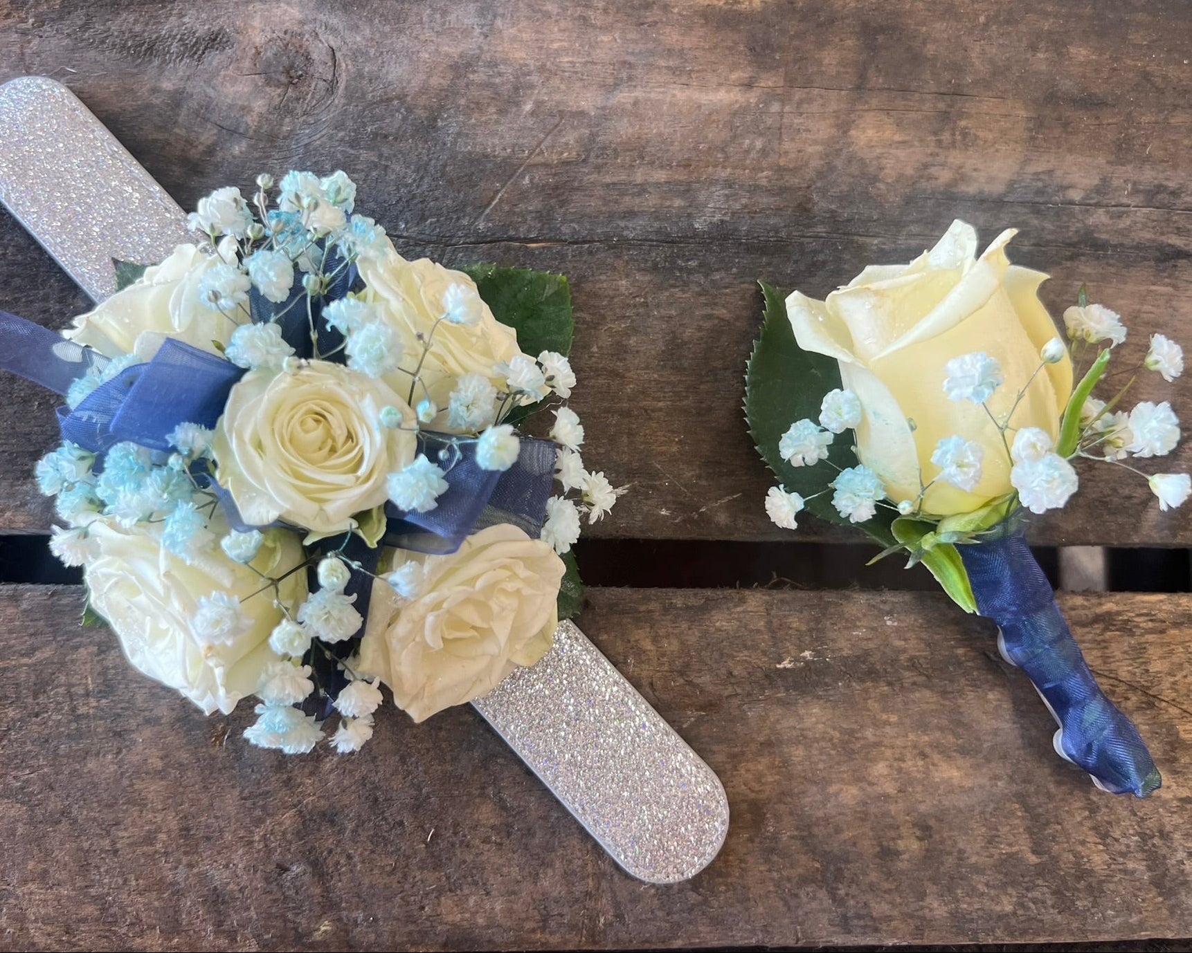 A matching floral wrist corsage and matching boutonniere set, featuring pale yellow roses, light blue baby's breath, and a blue ribbon, arranged on a glittery silver cross-shaped base over a rustic wooden background.