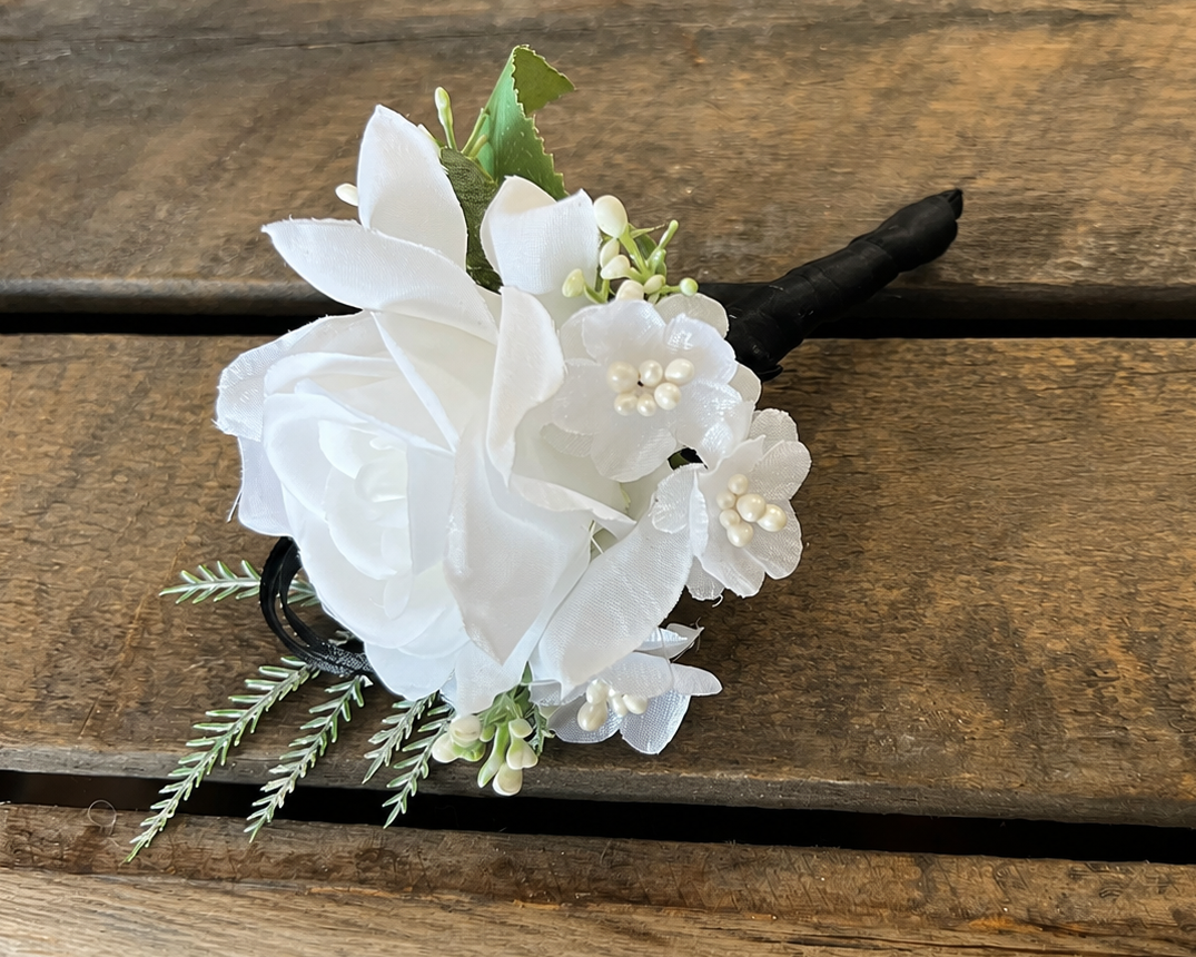 A white floral boutonniere with white artificial flowers, small green leaves, and a black stem, displayed on a rustic brown wooden surface.