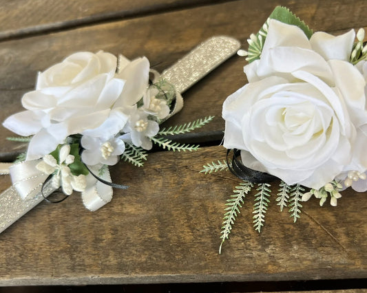 White faux rose wrist corsages with green and white filler flowers and ribbons, displayed on a rustic wooden background.