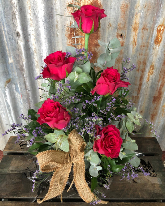 Arrangement of red roses with greenery and a burlap bow against a rustic metal background